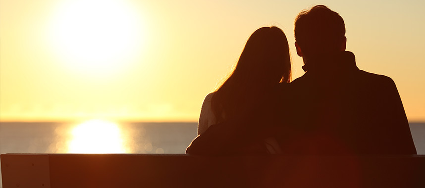 A young couple sit closely by the beach, watching the sunset as they think about their future retirement